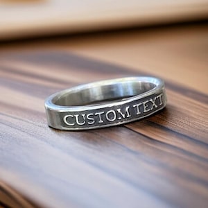May include: A silver ring with the engraved text "CUSTOM TEXT" is displayed on a wooden surface. The ring has a flat, rectangular profile with the text in a sans-serif font. The ring is the focal point, with the wooden surface providing a warm, neutral backdrop.