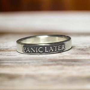 May include: A silver ring with the words "PANIC LATER" engraved on the band. The ring is set against a wooden surface, with the focus on the ring's inscription. The ring is a simple band style.