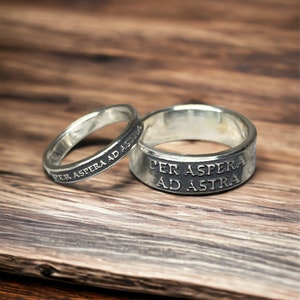 May include: Two silver rings with the inscription "Per Aspera Ad Astra" on a wooden surface.
