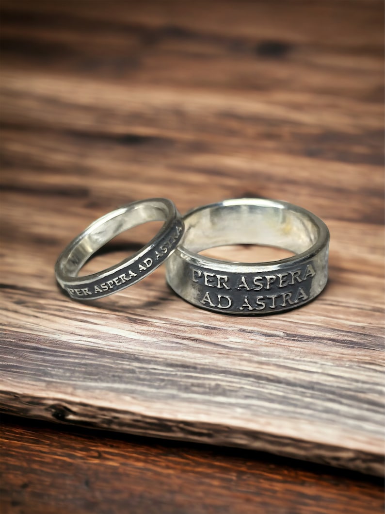 May include: Two silver rings with the inscription "Per Aspera Ad Astra" on a wooden surface.
