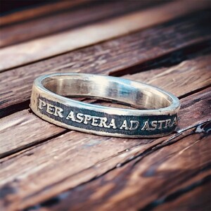 May include: A silver ring with the inscription "Per aspera ad astra" on a wooden surface.