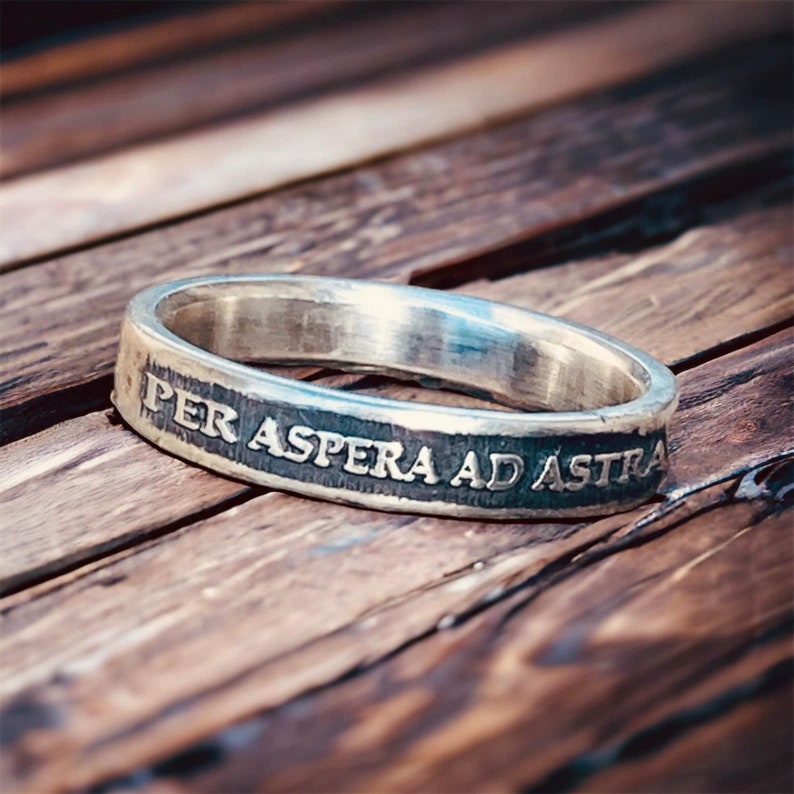 May include: A silver ring with the inscription "Per aspera ad astra" on a wooden surface.