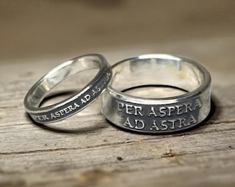 May include: Two silver rings with a polished finish, each featuring the engraved Latin phrase PER ASPERA AD ASTRA. The text is darkened to stand out against the metallic surface. The rings have a smooth, rounded band design with a consistent width.