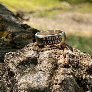 May include: A silver ring with the inscription "Per aspera ad astra" on a rough, brown, textured surface.