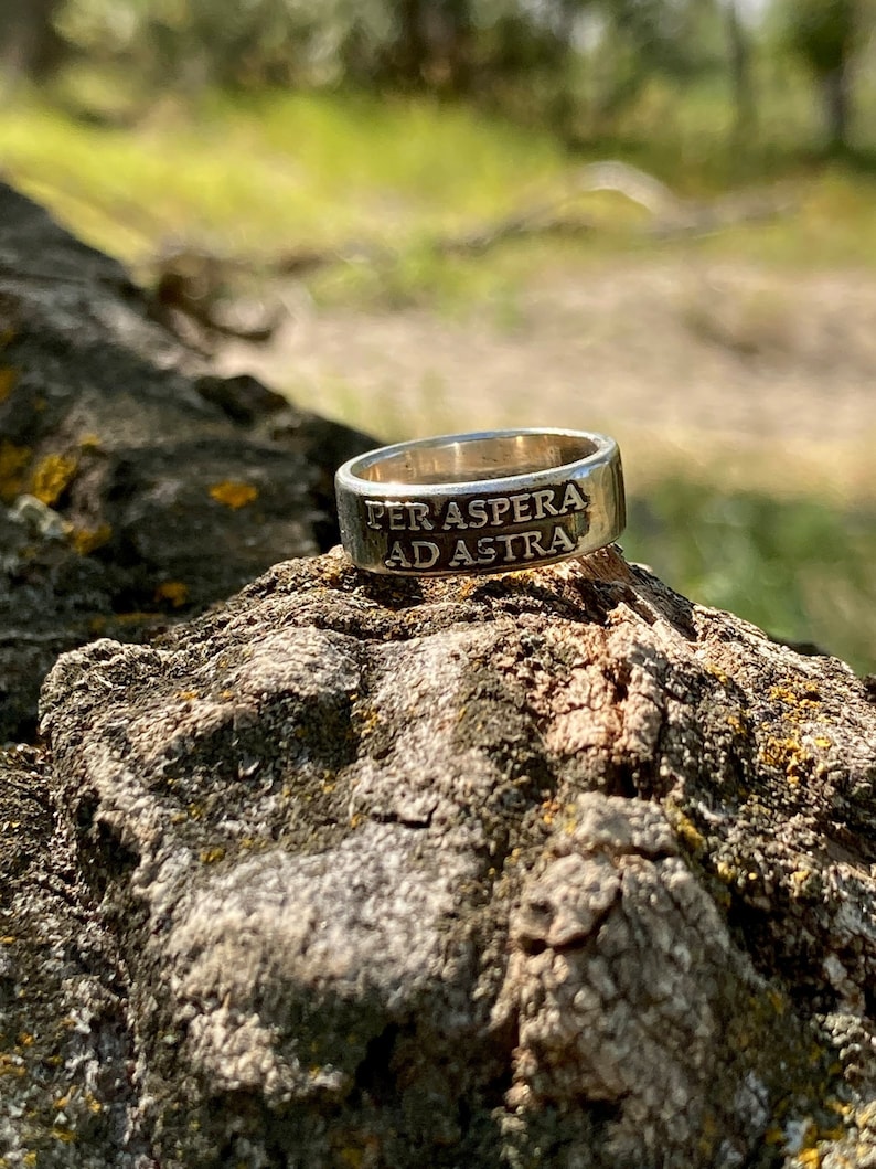 May include: A silver ring with the inscription "Per aspera ad astra" on a rough, brown, textured surface.