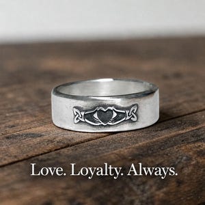 May include: A silver band ring featuring an engraved Claddagh design with two hands holding a heart and a crown, accompanied by the text Love. Loyalty. Always. The metal has a polished finish with darkened recessed details in the engraving.