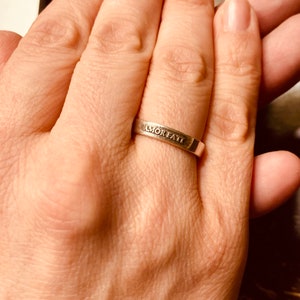May include: A silver-toned metal band ring featuring the engraved text AMOR FATI on the outer surface. The ring has a smooth, flat profile and a polished finish, encircling the finger with a simple, uniform width throughout the entire band.