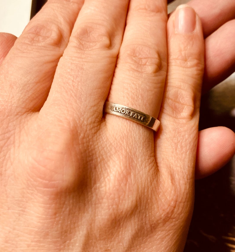 May include: A silver-toned metal band ring featuring the engraved text AMOR FATI on the outer surface. The ring has a smooth, flat profile and a polished finish, encircling the finger with a simple, uniform width throughout the entire band.