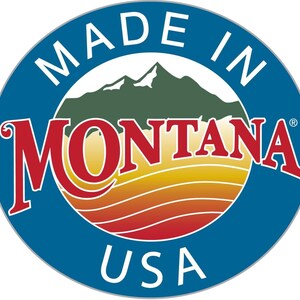 May include: A circular emblem featuring a blue border with the white text MADE IN USA. The center displays a stylized mountain range in green above a sunset landscape with horizontal orange and yellow stripes, and the red text MONTANA.