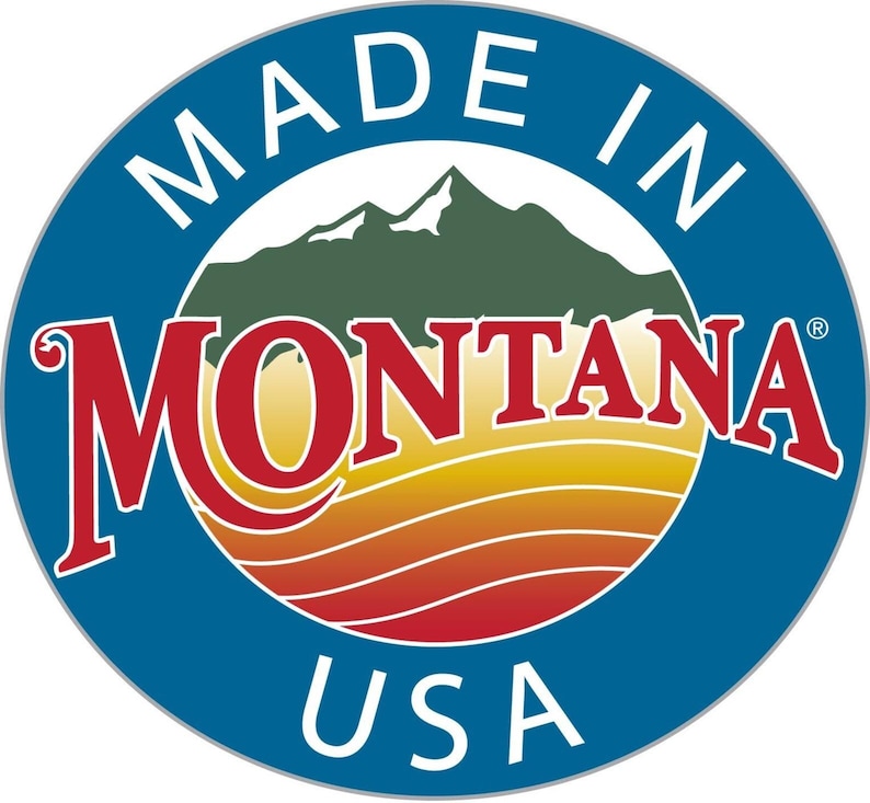 May include: A circular emblem featuring a blue border with the white text MADE IN USA. The center displays a stylized mountain range in green above a sunset landscape with horizontal orange and yellow stripes, and the red text MONTANA.