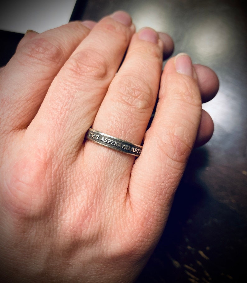 May include: A silver ring with the inscription "Per Aspera Ad Astra" on a person's finger.