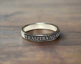 May include: A silver-toned metal ring featuring a smooth, polished finish. The outer surface of the band is engraved with the Latin phrase PER ASPERA AD ASTRA in clear, capital letters. The ring has a simple, uniform band design.