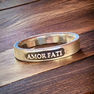 May include: A silver-colored metal ring with a smooth, polished finish. The outer surface of the band features a rectangular, dark-toned recessed area with the words AMOR FATI engraved in capital letters across the center of the band.