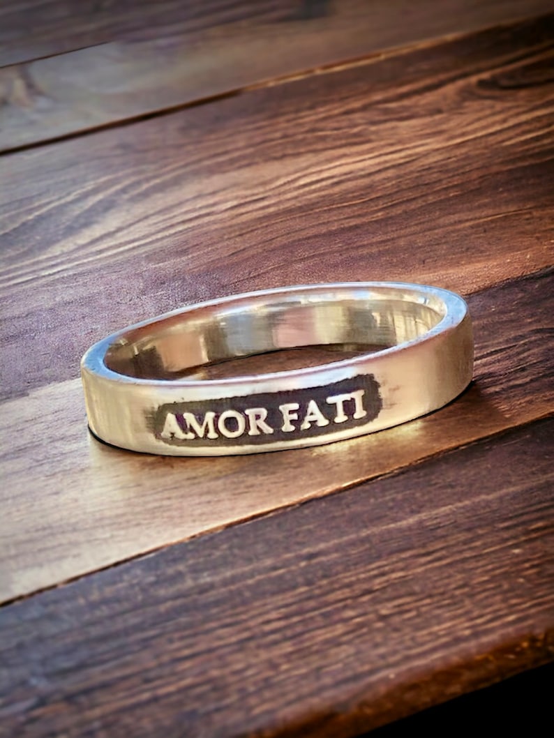 May include: A silver-colored metal ring with a smooth, polished finish. The outer surface of the band features a rectangular, dark-toned recessed area with the words AMOR FATI engraved in capital letters across the center of the band.
