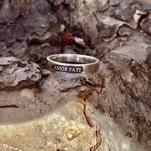 May include: A silver-toned metal ring featuring a flat band with the words AMOR FATI engraved in a recessed, darkened rectangular panel on the front surface. The ring has a smooth, polished finish and a consistent width around the entire band.
