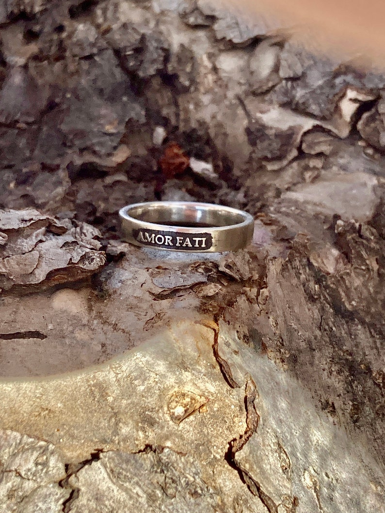 May include: A silver-toned metal ring featuring a flat band with the words AMOR FATI engraved in a recessed, darkened rectangular panel on the front surface. The ring has a smooth, polished finish and a consistent width around the entire band.