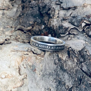 May include: A silver ring with the inscription "Per Aspera Ad Astra" on a rough, gray bark background.