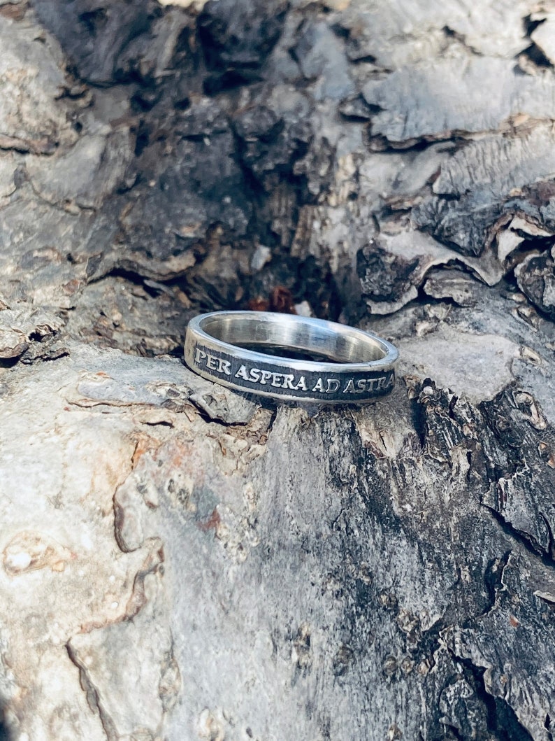 May include: A silver ring with the inscription "Per Aspera Ad Astra" on a rough, gray bark background.