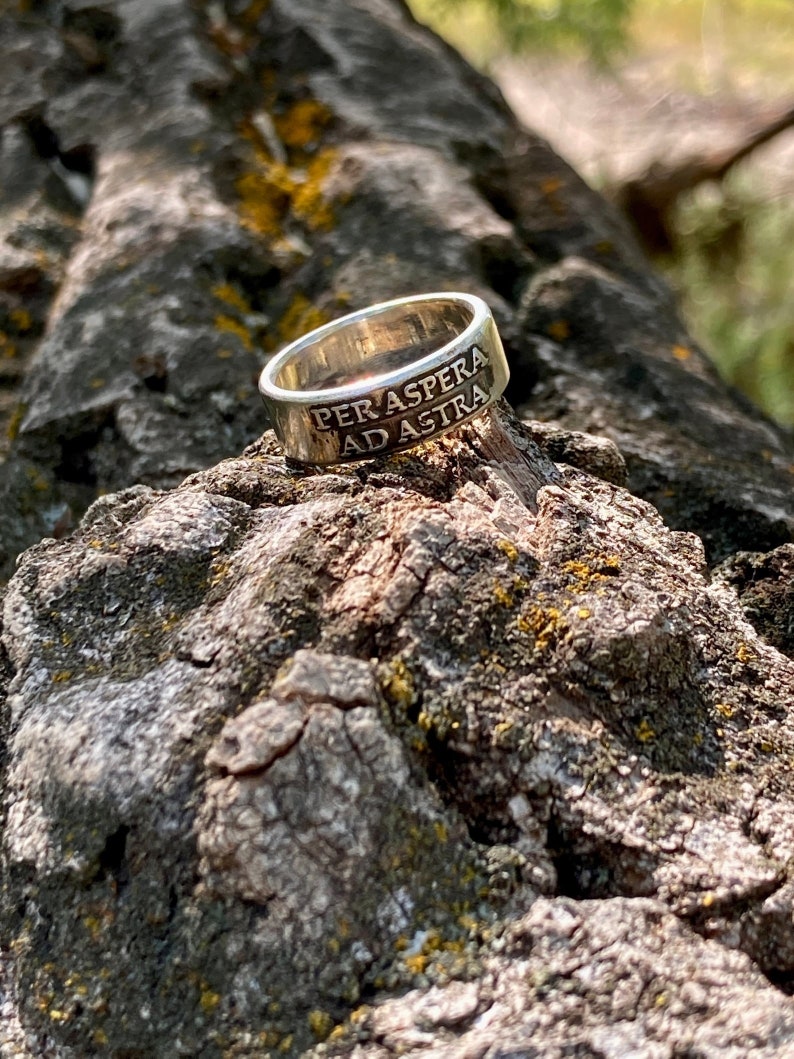 May include: A silver ring with the inscription "Per aspera ad astra" on a tree branch.