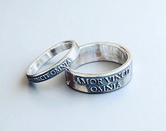 May include: Two silver-toned rings with a polished finish. The thinner ring features the engraved text AMOR VINCIT OMNIA. The wider ring also displays the engraved text AMOR VINCIT OMNIA in a clear, capitalized font around the outer band surface.