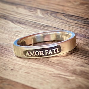 May include: A silver-toned metal ring featuring a flat, rectangular front panel with the words AMOR FATI engraved in capital letters. The engraving is filled with a dark, contrasting color, and the band has a smooth, polished finish throughout.