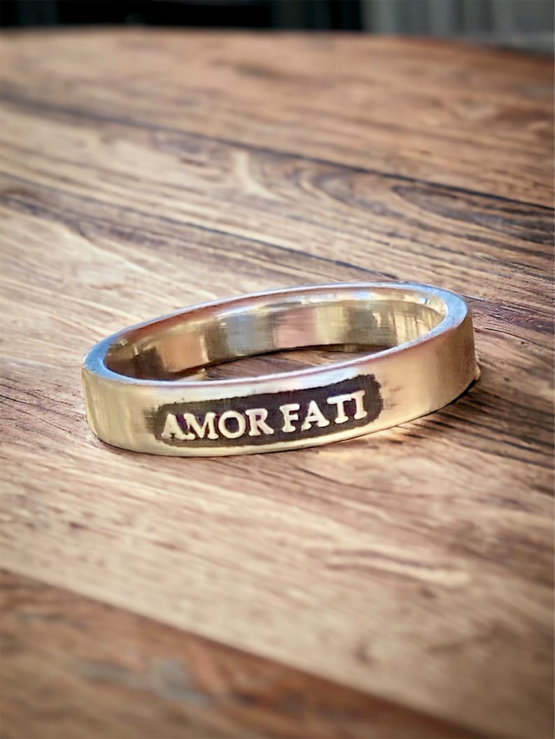 May include: A silver-toned metal ring featuring a flat, rectangular front panel with the words AMOR FATI engraved in capital letters. The engraving is filled with a dark, contrasting color, and the band has a smooth, polished finish throughout.