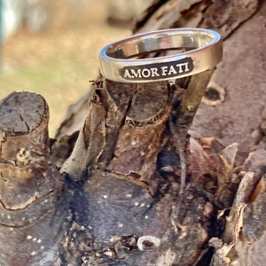 May include: A silver-toned metal ring with a smooth, polished finish. The outer surface of the band features the engraved text AMOR FATI in capital letters, with the lettering filled in with a dark, contrasting color for clear visibility.
