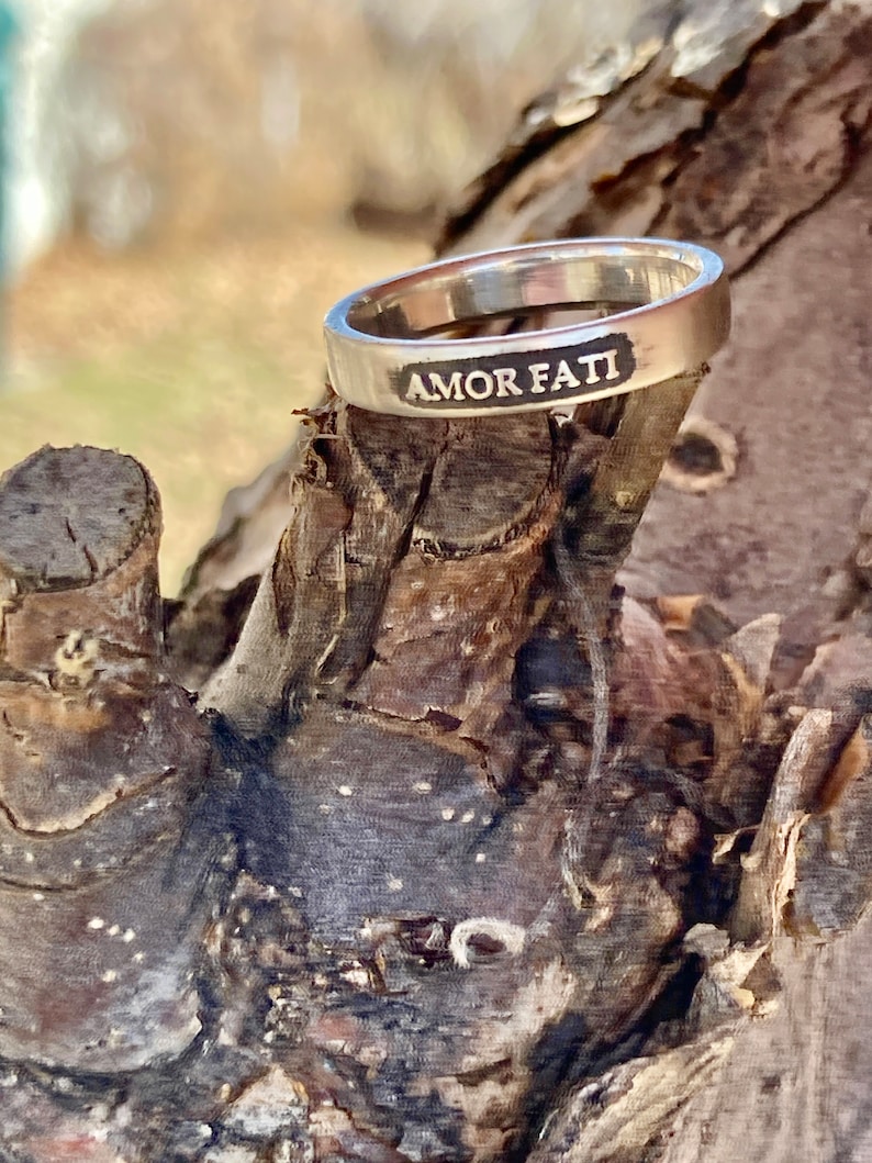 May include: A silver-toned metal ring with a smooth, polished finish. The outer surface of the band features the engraved text AMOR FATI in capital letters, with the lettering filled in with a dark, contrasting color for clear visibility.