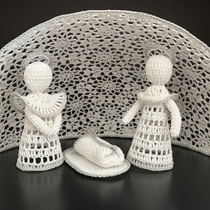 May include: A handcrafted, white crochet nativity scene. The scene includes two angel figures, a cradle, and a decorative backdrop. The angels have halos and are dressed in white crochet garments. The backdrop is a large, semi-circular piece with a floral pattern.