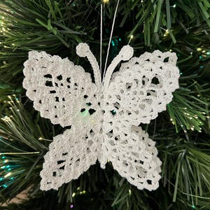 May include: A white crocheted butterfly ornament featuring an intricate openwork lace pattern with four distinct wings and two antennae, suspended by a thin white string. The butterfly is crafted from fine white thread with a delicate texture.
