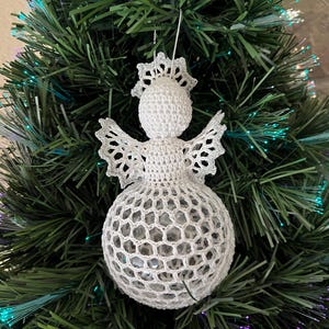 May include: A white crocheted angel Christmas ornament with a halo and openwork wings and skirt. The angel is hanging on a green Christmas tree with fiber optic lights. The ornament is handmade.