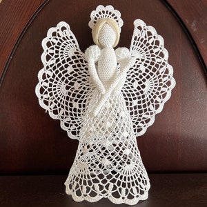 Puede incluir: Figurita de ángel de crochet blanco con halo y alas. El ángel está de pie sobre una superficie de madera.