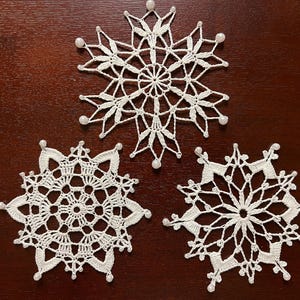 May include: Three white crocheted snowflakes with pearl beads on a brown wooden surface.