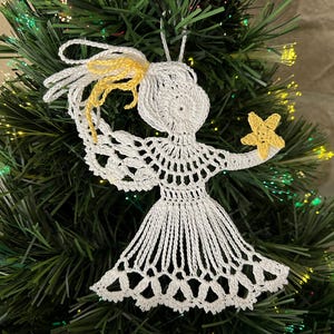 May include: A handmade white crochet angel Christmas ornament with yellow accents, holding a yellow star. The angel is hanging on a green Christmas tree with lights.