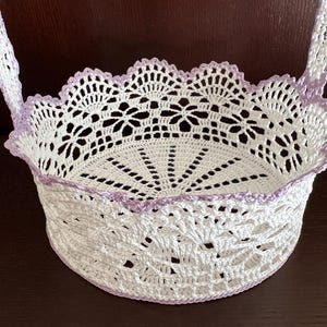 Peut inclure: Un panier blanc crocheté avec un bord festonné et une poignée. Le panier présente un motif floral délicat et est bordé d'une bordure lilas clair. Le design ajouré permet de voir le contenu.