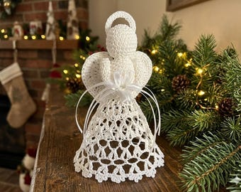 May include: A white crocheted angel figurine featuring a circular halo, rounded head, and wide wings. The body is crafted with an openwork lace pattern, and a white satin ribbon is tied into a bow at the center, accentuating the waist area.