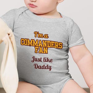 May include: A grey baby onesie with the text "I'm a Commanders Fan Just like Daddy" in yellow and maroon. The baby is wearing the onesie and looking at a stuffed animal.