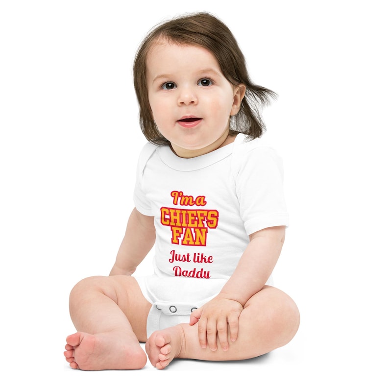May include: A white baby onesie with red and yellow text that reads "I'm a Chiefs Fan Just like Daddy". A baby is wearing the onesie and sitting on a white background.