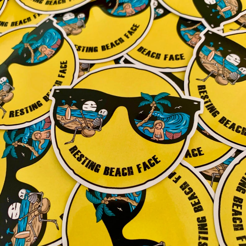 May include: A yellow sticker with a smiley face wearing sunglasses. The sunglasses reflect a beach scene with a palm tree, ocean waves, and a person laying on a beach towel. The text "RESTING BEACH FACE" is written in black letters on the sticker.