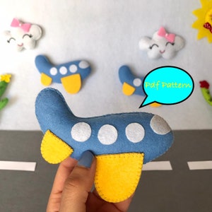 May include: A blue felt airplane with yellow wings and white circles. The airplane is on a gray felt road with white lines. There are two white felt clouds with pink bows and a yellow felt sun with a red bow. There is also a green felt cactus on each side of the road. The text "Pdf Pattern" is written in a blue speech bubble.