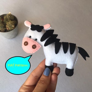 May include: A white and black felt zebra toy with pink ears and black eyes. The zebra has a black mane and tail. The toy is being held by a hand. The text "Pdf Pattern" is in a blue speech bubble.