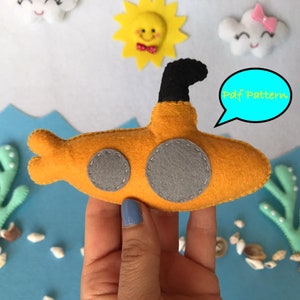 May include: A yellow felt submarine with a black periscope and gray portholes. The submarine is held in a hand against a blue background with white clouds, a yellow sun, and green seaweed.