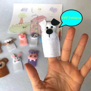 May include: A set of felt finger puppets featuring a lion, sheep, pig, cat, dog, rabbit, and hippopotamus. The puppets are colorful and have friendly faces. The image also includes a hand wearing a dog finger puppet and a speech bubble that says "Pdf Pattern".