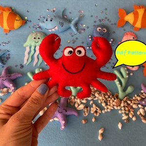 May include: A red felt crab with a smiling face and googly eyes. The crab is surrounded by other felt sea creatures, including a blue shark, a purple starfish, and a yellow fish. The text "Pdf Pattern" is written in a speech bubble.