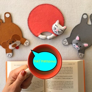 May include: Three felt animal-shaped coasters, a brown dog, a gray cat, and a white cat with red accents. The coasters are on a white surface with a book and a cup of coffee. The text "Pdf Patterns" is on the cup.