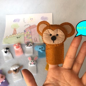May include: A set of felt finger puppets featuring various animals, including a lion, dog, pig, cat, elephant, rabbit, sheep, and a hippopotamus. The puppets are all white with different coloured facial features. The puppets are arranged on a white surface. A brown bear finger puppet is on a hand with a blue speech bubble that says "Pdf Pattern".