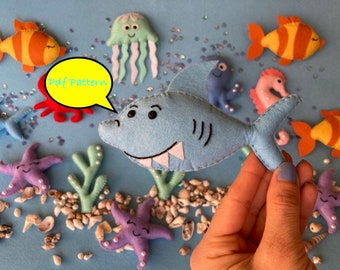 May include: A blue felt shark with white teeth and black eyes. The shark is surrounded by felt sea creatures, including a starfish, a jellyfish, a seahorse, and an octopus. The text "Pdf Pattern" is written in a speech bubble.