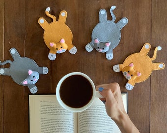 May include: Four felt cat coasters, two orange and two gray, with pink noses and white paws, surrounding a cup of coffee on a book.