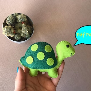 May include: Green felt turtle with yellow spots, a sewn smile, and black eyes. The turtle is being held in a hand. A blue speech bubble with the text "Pdf Pattern" is above the turtle.