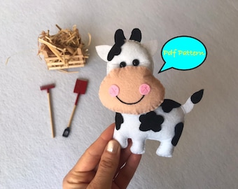 Peut inclure: Une vache en feutre blanche et noire avec des joues roses et des yeux noirs. La vache tient une bulle de dialogue qui dit "Pdf Pattern".